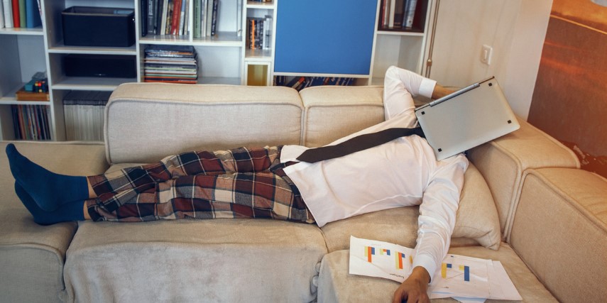 A man lies on a couch with a laptop over his face and papers with graphs under his arm.