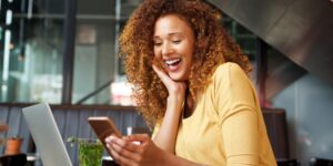 A woman in a yellow shirt sits at a desk, smiling at something she sees on her phone screen.