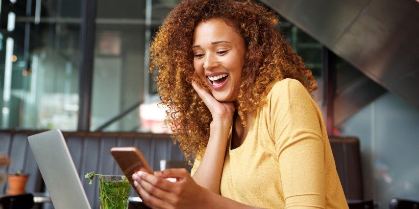A woman in a yellow shirt sits at a desk, smiling at something she sees on her phone screen.