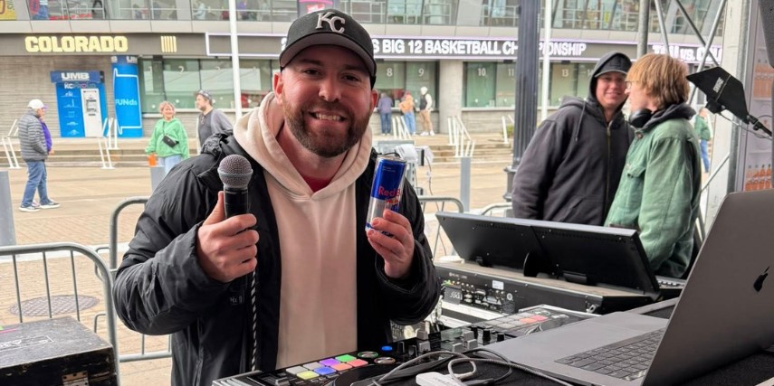 A DJ stands in front of electronic equipment with a microphone in one hand and a can of energy drink in the other.