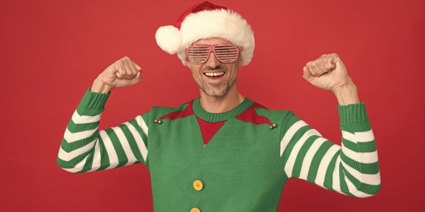 A man dressed as an elf stands in front of a red background and raises his fists in a celebratory manner.
