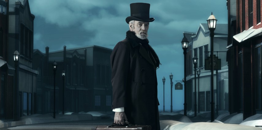 A man dressed as Ebenezer Scrooge turns to look to his right. He stands on a dark street.