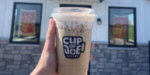 Close-up of a latte in a to-go cup with the logo of "Cup of Joe" on the front.