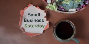 A piece of cardboard with ripped edges says, "Small Business Saturday," next to a cup of coffee and some succulent potted plants.