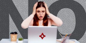 A woman sits at a desk with the word "No" across the backdrop. She's holding her hands to her head in frustration and has an overwhelmed look on her face.