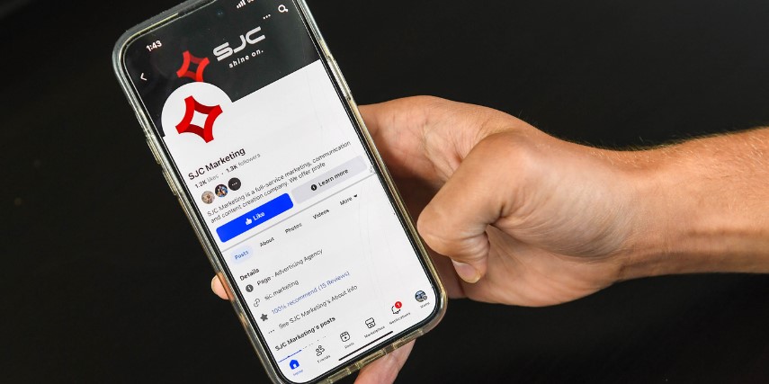 A hand holds a smartphone featuring the social media profile page for SJC Marketing.
