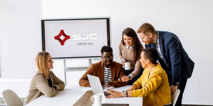 A group of people gather around a laptop in an office and a screen with "SJC" is behind them.