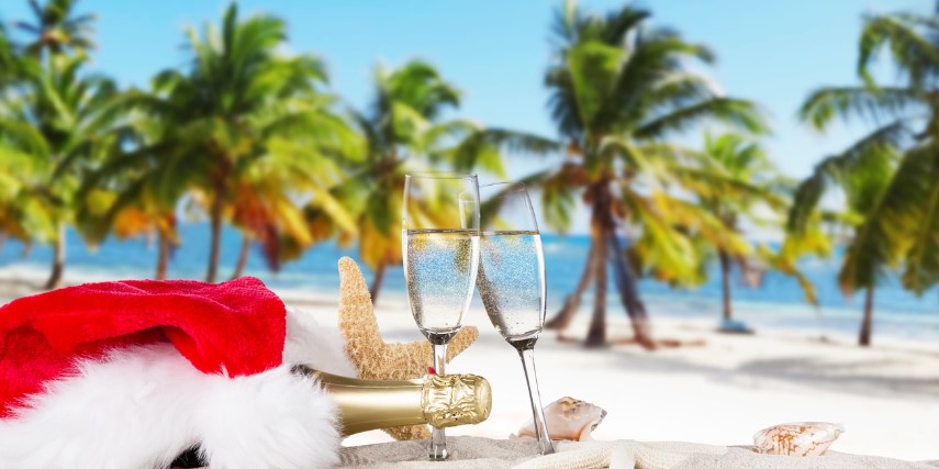 Two glasses of champagne and a Santa hat sit on the sand in front of palm trees on the beach. 