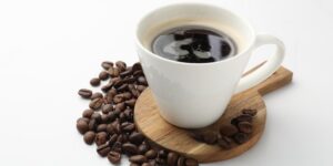 A black cup of coffee rests on a small wood paddle with coffee beans scattered near it.