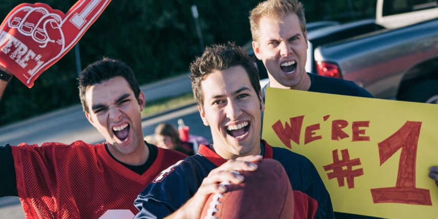 Three football fans shout, hold a sign and hold up a foam finger celebrating their team.