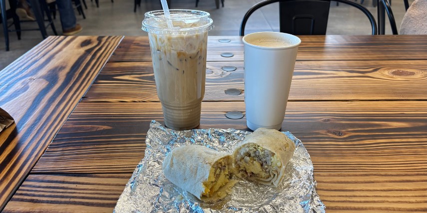 An iced coffee, hot coffee and a breakfast wrap on a wood table in a coffee shop.