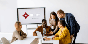 A group of people gather over a table strategizing with the SJC logo on a monitor behind them.