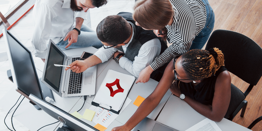A group of coworkers strategize at a table, with some leaning over those seated as they discuss strategy.