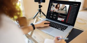 A woman sits at a desk looking at a laptop with a smartphone on a tripod near the laptop.