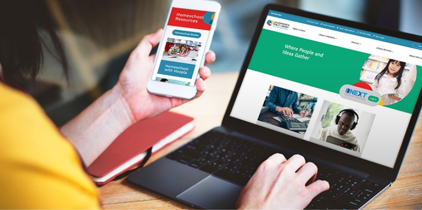 A person sits at a desk holding a smartphone as they look at a laptop website.