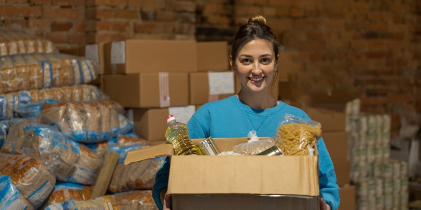 A woman holds a cardboard box filled with a bottle of canola oil, a bag of pasta and other food items.
