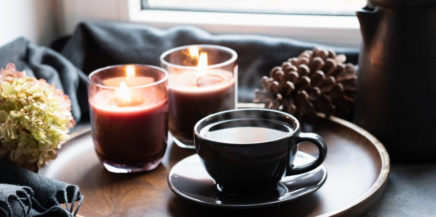 Two candles and a cup of coffee sit on a wooden tray near a pinecone, hydrangea bloom and a blanket.