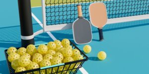 A basket of balls and two pickleball paddles lean against a pickleball court net.