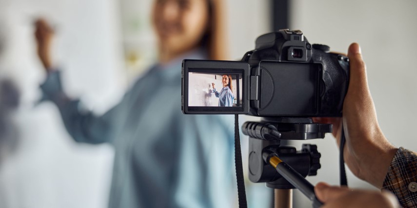 A video camera filming a woman standing at a whiteboard giving a tutorial.