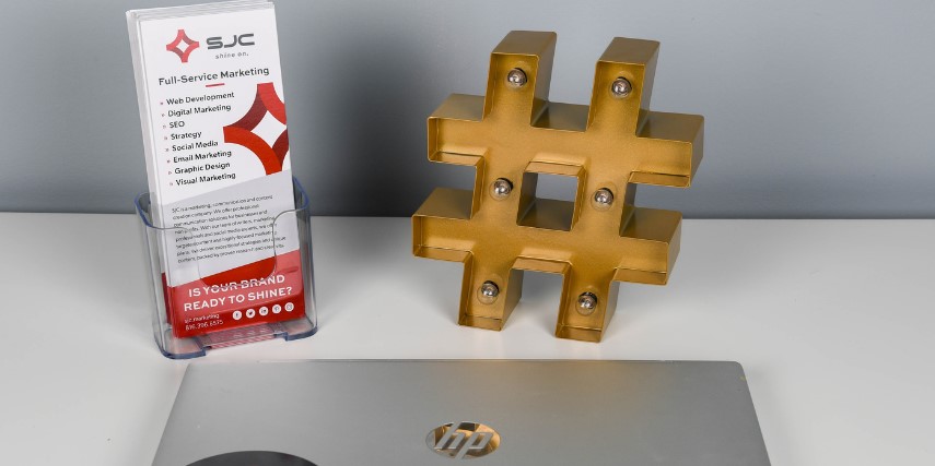 A laptop, rack card and a lighted hashtag sign on a white desk.