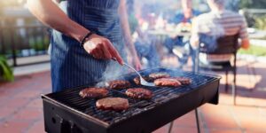 A person stands at a grill flipping hamburgers with a gathering of people in the background.