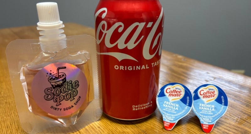 A packet of dirty soda flavoring, a Coke and two flavored creamers on a table.