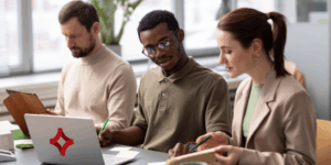 Three people sit at a long desk discussing a strategy. An SJC logo is featured on the laptop on the desk.