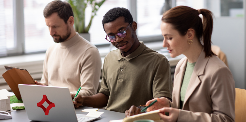 Three people sit at a long desk discussing a strategy. An SJC logo is featured on the laptop on the desk.