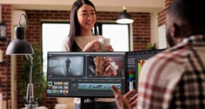 A woman stands talking to a man editing video content on a large monitor.