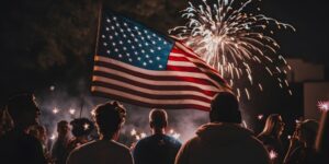 A group of people watches a fireworks display with a flag in the foreground.
