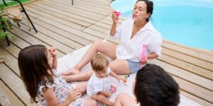 A woman sits with three young children on a deck blowing bubbles near a pool.