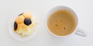 A piece of cake with a soccer ball design is next to a cup of coffee with cream on a white table.