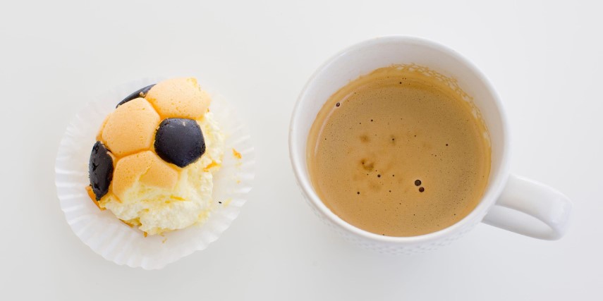 A piece of cake with a soccer ball design is next to a cup of coffee with cream on a white table.