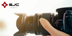 A woman's hand holds the lens of a professional camera. The SJC logo is in the corner of the image.
