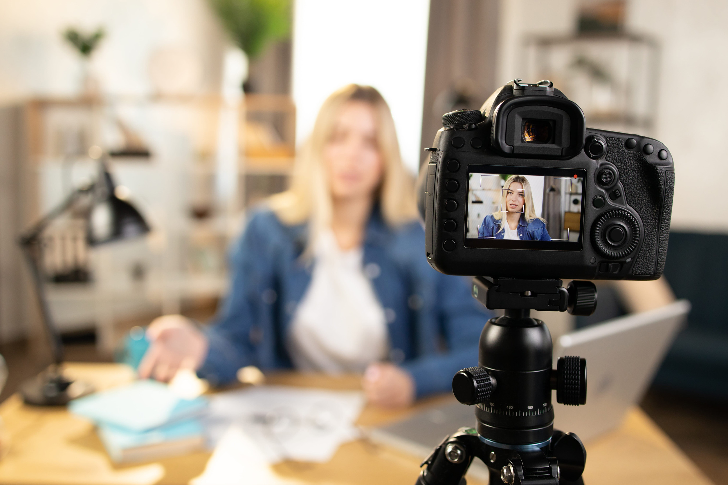 Earned Media Blond businesswoman in casual clothes filming educational lecture on video camera while sitting at table. Focus on camera screen. Blur background.