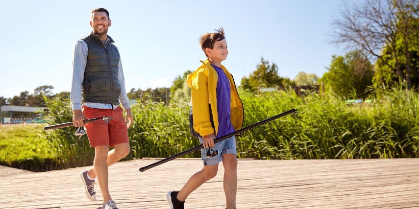 A man and a young teenager walk near a river with fishing poles.