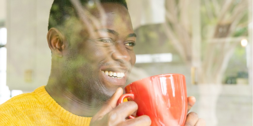 Reflection of a man standing at his window drinking coffee from a large red mug.