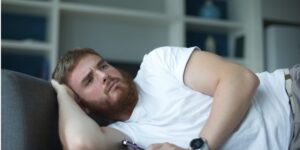 A man lies on a couch with a remote control in his hand, looking concerned.
