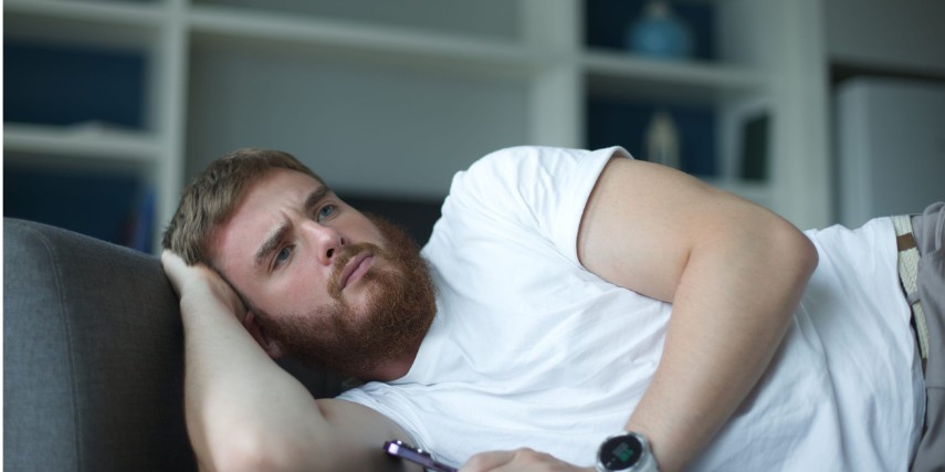A man lies on a couch with a remote control in his hand, looking concerned.