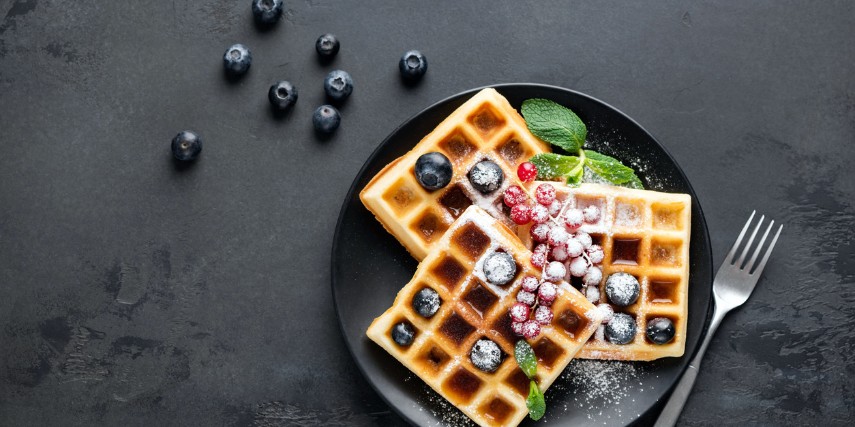 A plate of waffles with berries and powdered sugar on a gray-topped table with blueberries scattered nearby.