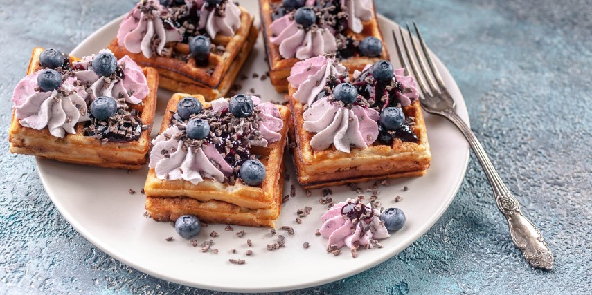 Waffles decorated with purple whipped cream and blueberries on a white plate with a fork perched on the side.