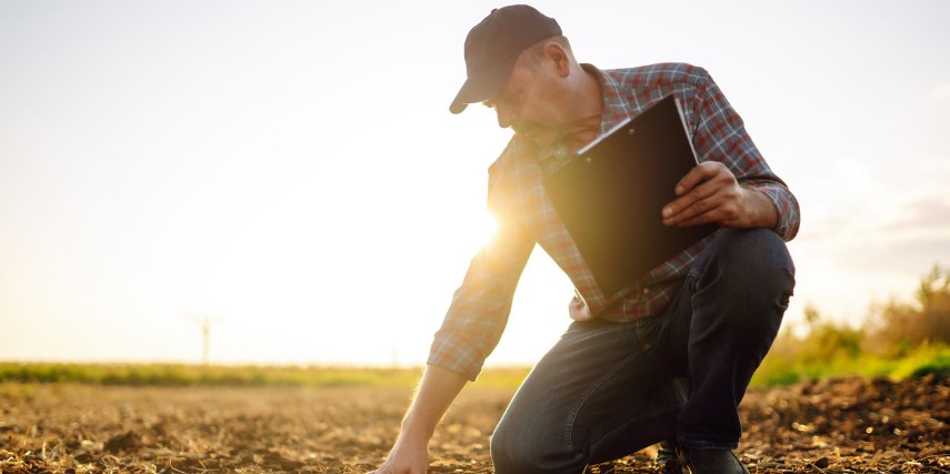 A farmer kneels on the ground holding a clipboard as he assesses the soil.
