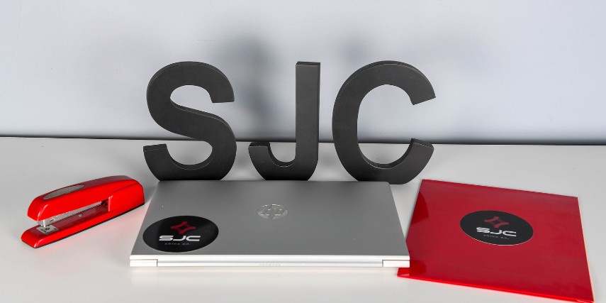 Large black "SJC" behind a laptop and between a red stapler and a red folder.