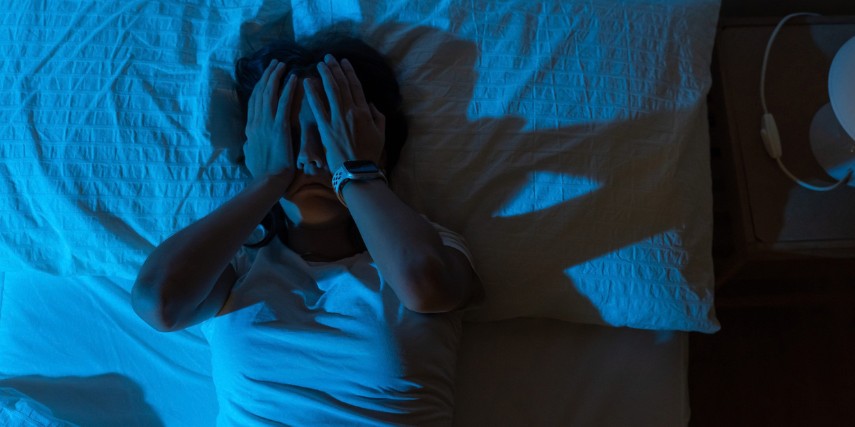 A woman lies in bed in a darkened room with her hands covering her face.