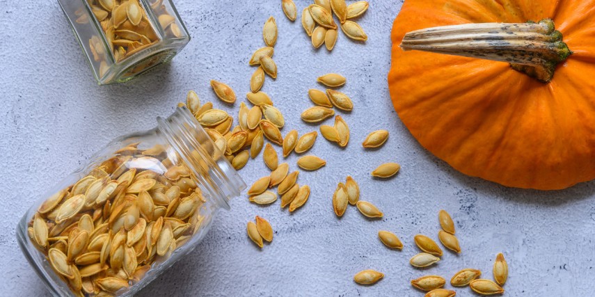 Seeds scattered on a table next to two jars and a pumpkin.